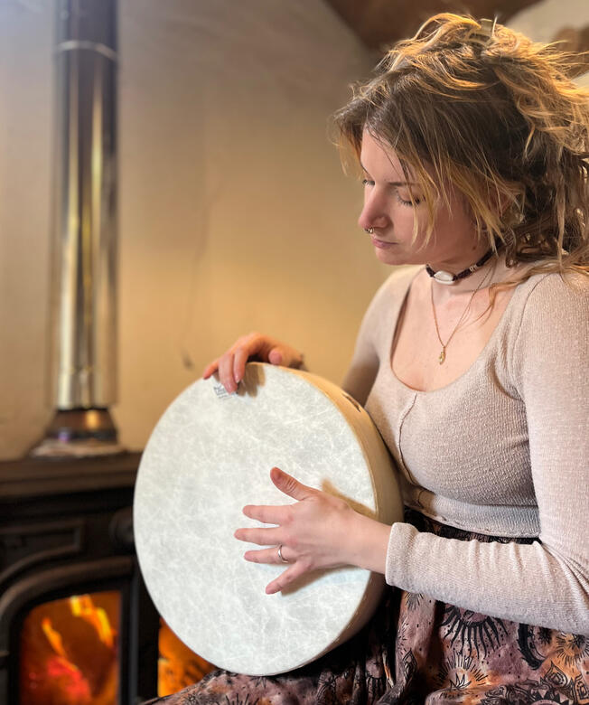 Image of Carli, birth doula, playing a hand drum by a fireplace